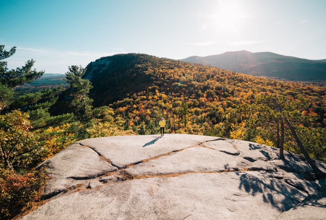 A person standing on a large rock outcropping looking out at fall leaves on the mountains in the distance. 